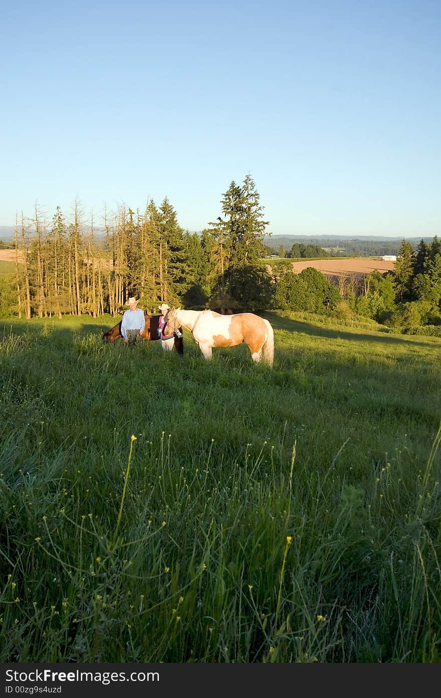 Two farmers smiling for the camera in front of trees as their horses are near them on a grassy hill. - vertically framed. Two farmers smiling for the camera in front of trees as their horses are near them on a grassy hill. - vertically framed