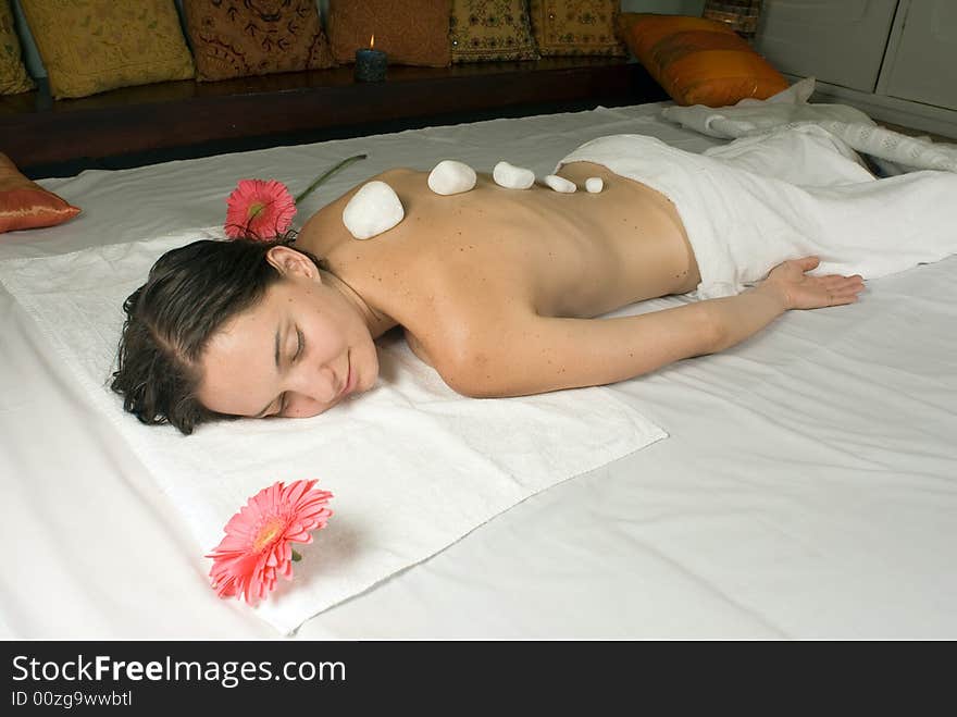 A young girl lying down on a white towel with pink flowers near her, hot rocks are on her back. - horizontally framed. A young girl lying down on a white towel with pink flowers near her, hot rocks are on her back. - horizontally framed