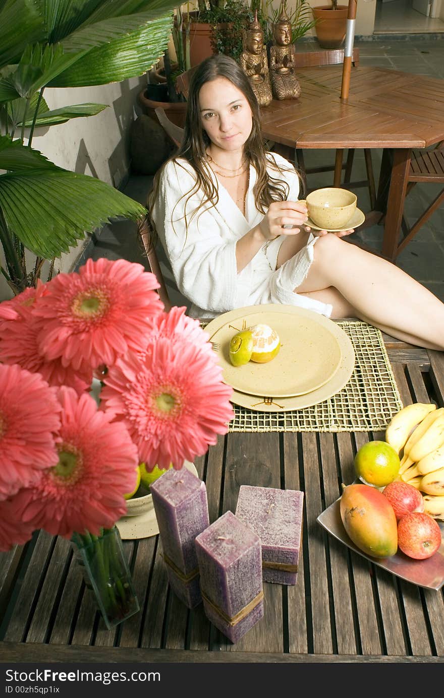 A young girl sitting at a table with plants, fruits, and flowers on it. She is holding a cup while looking at the camera. - vertically framed. A young girl sitting at a table with plants, fruits, and flowers on it. She is holding a cup while looking at the camera. - vertically framed