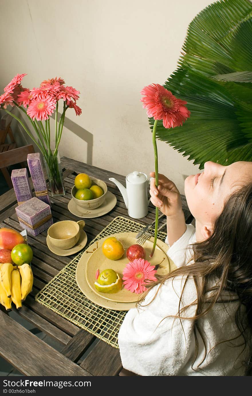 An attractive young girl, smelling a pink flower, sitting at a table, while fruits and plants surround her. - vertically framed. An attractive young girl, smelling a pink flower, sitting at a table, while fruits and plants surround her. - vertically framed