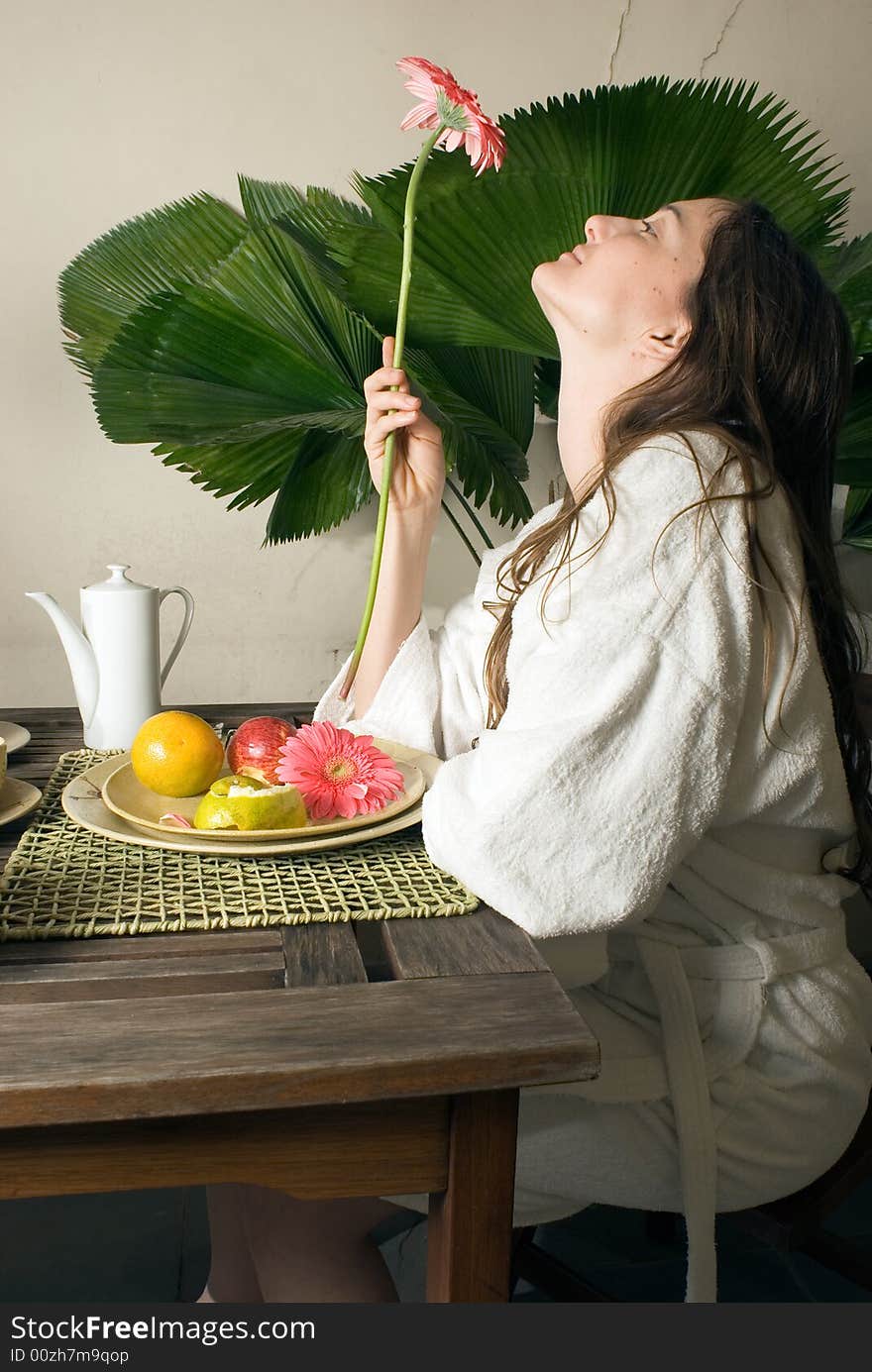 An attractive young girl outside, staring at a pink flower, sitting at a table, while fruits and plants surround her. - vertically framed. An attractive young girl outside, staring at a pink flower, sitting at a table, while fruits and plants surround her. - vertically framed