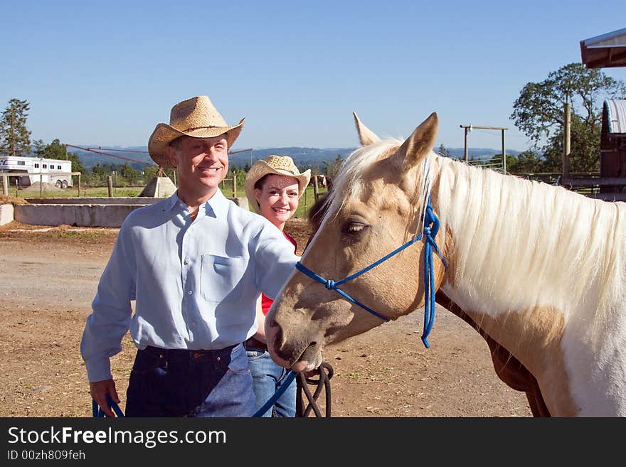 A Cowgirl and Cowboy smiling as they pet a blonde horse. Hoizontally framed photograph. A Cowgirl and Cowboy smiling as they pet a blonde horse. Hoizontally framed photograph