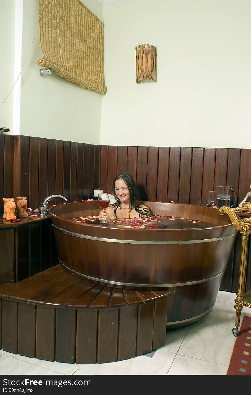 Young lady smiling at a wine glass, in a bathtub, roses in water, candles surrounding. - vertically framed. Young lady smiling at a wine glass, in a bathtub, roses in water, candles surrounding. - vertically framed