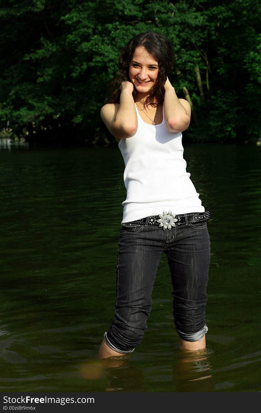 The girl in dark blue jeans and a white vest costs in water on a background of green trees. The girl in dark blue jeans and a white vest costs in water on a background of green trees