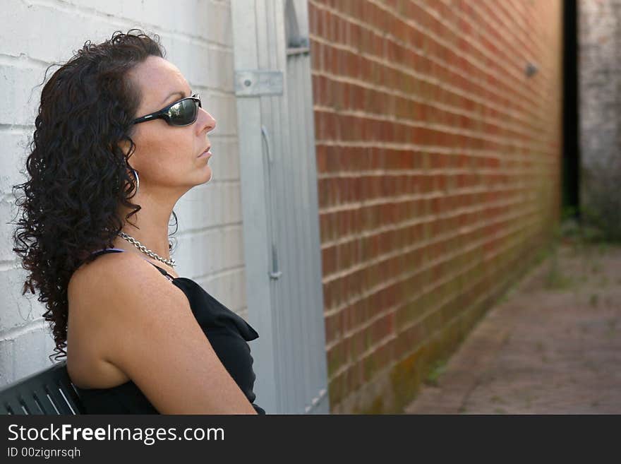 A pretty woman sitting outside of a long empty alley near a red brick wall.