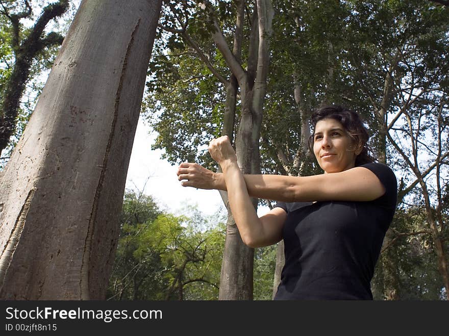 A woman is standing in the park. She is stretching her arms across her chest. Horizontally framed photo. A woman is standing in the park. She is stretching her arms across her chest. Horizontally framed photo.