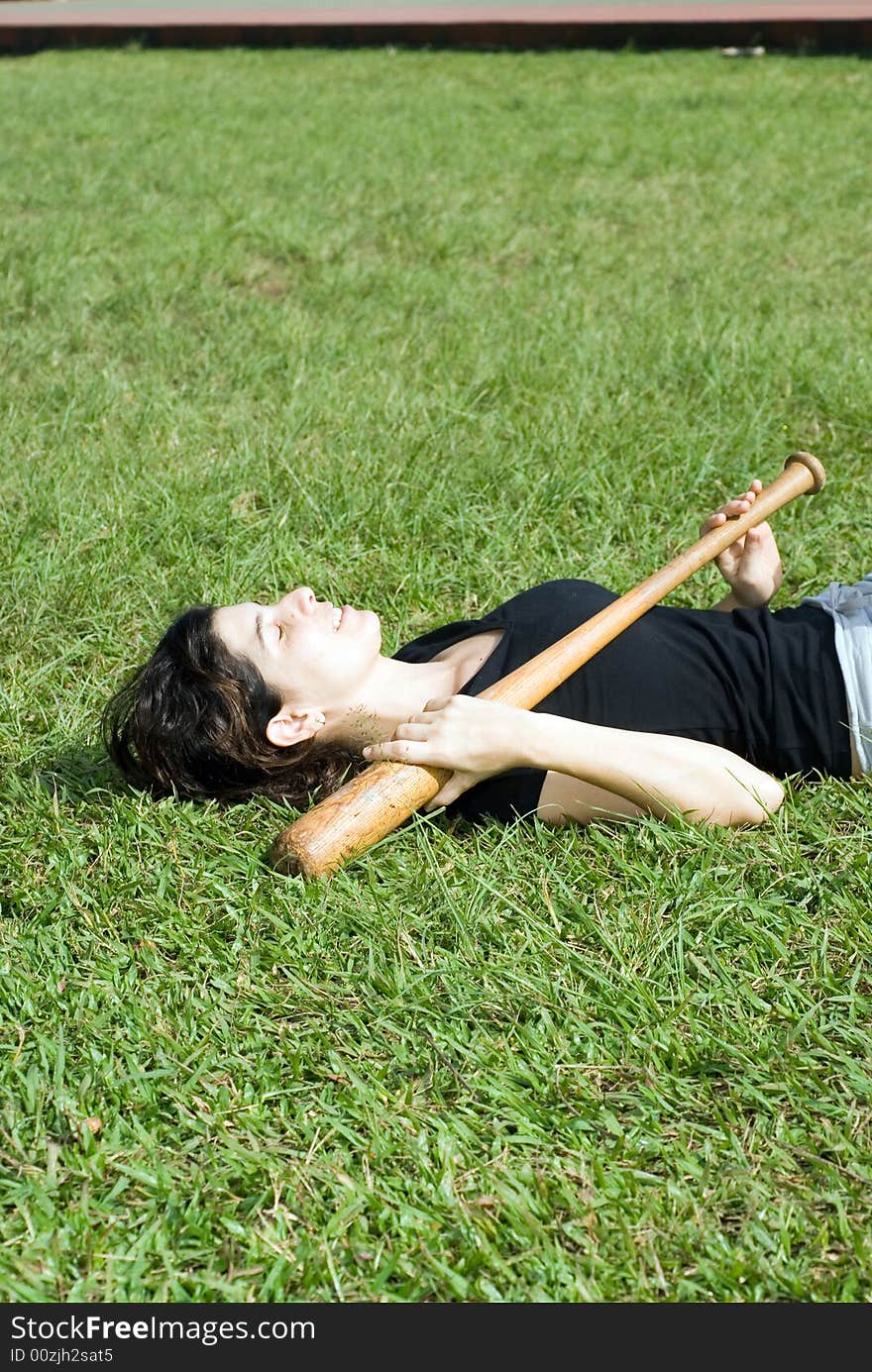 A woman is laying on the grass at the park. She is holding a baseball bat across her chest, and appears to be sleeping or resting. Vertically framed shot. A woman is laying on the grass at the park. She is holding a baseball bat across her chest, and appears to be sleeping or resting. Vertically framed shot.