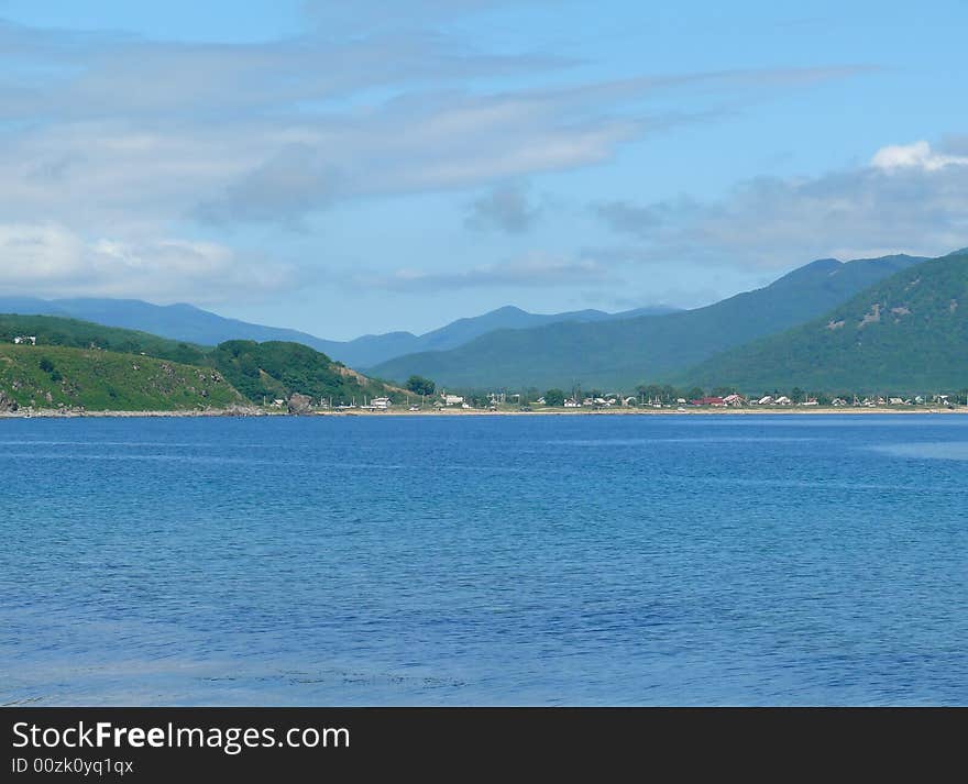 A landscape at sea. A blue seawater on foreground. A sky with white clouds, green mountaines and village at bank. Russian Far East, Primorye, Sokolovka village. A landscape at sea. A blue seawater on foreground. A sky with white clouds, green mountaines and village at bank. Russian Far East, Primorye, Sokolovka village.