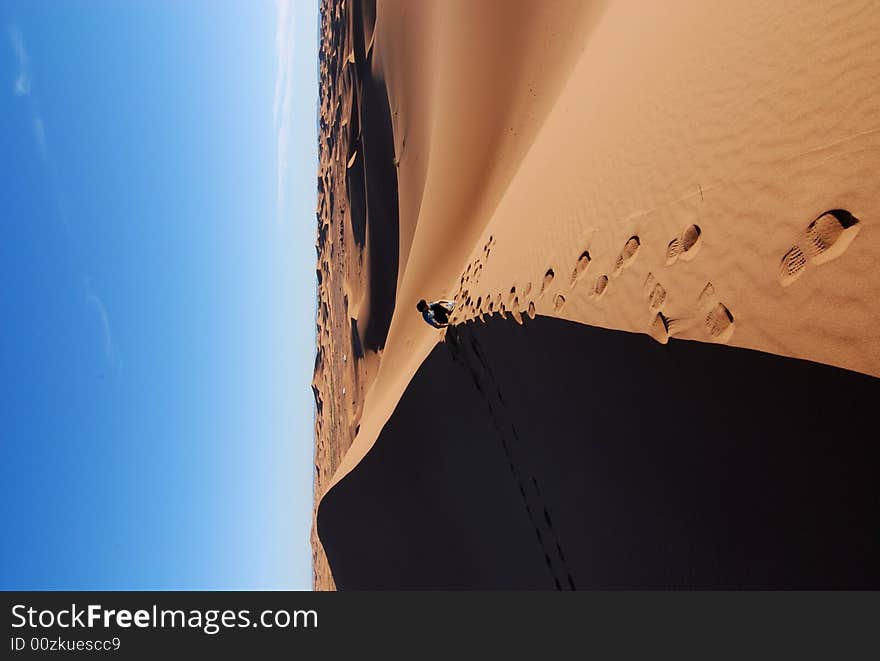 A man sits on a sand dune in Morocco in the Sahara desert. His footprints are seen in the sand. A man sits on a sand dune in Morocco in the Sahara desert. His footprints are seen in the sand.