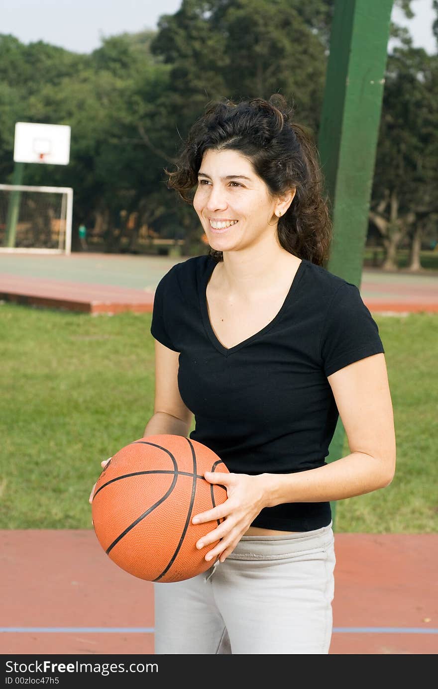 Young, attractive, happy woman is standing on an outdoor basketball court.  She is holding a basketball and looking in the other direction.  Vertically framed shot. Young, attractive, happy woman is standing on an outdoor basketball court.  She is holding a basketball and looking in the other direction.  Vertically framed shot.