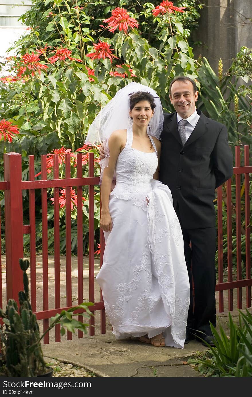 Married couple pose in front of a red fence, with red flowers in background. - vertically framed. Married couple pose in front of a red fence, with red flowers in background. - vertically framed