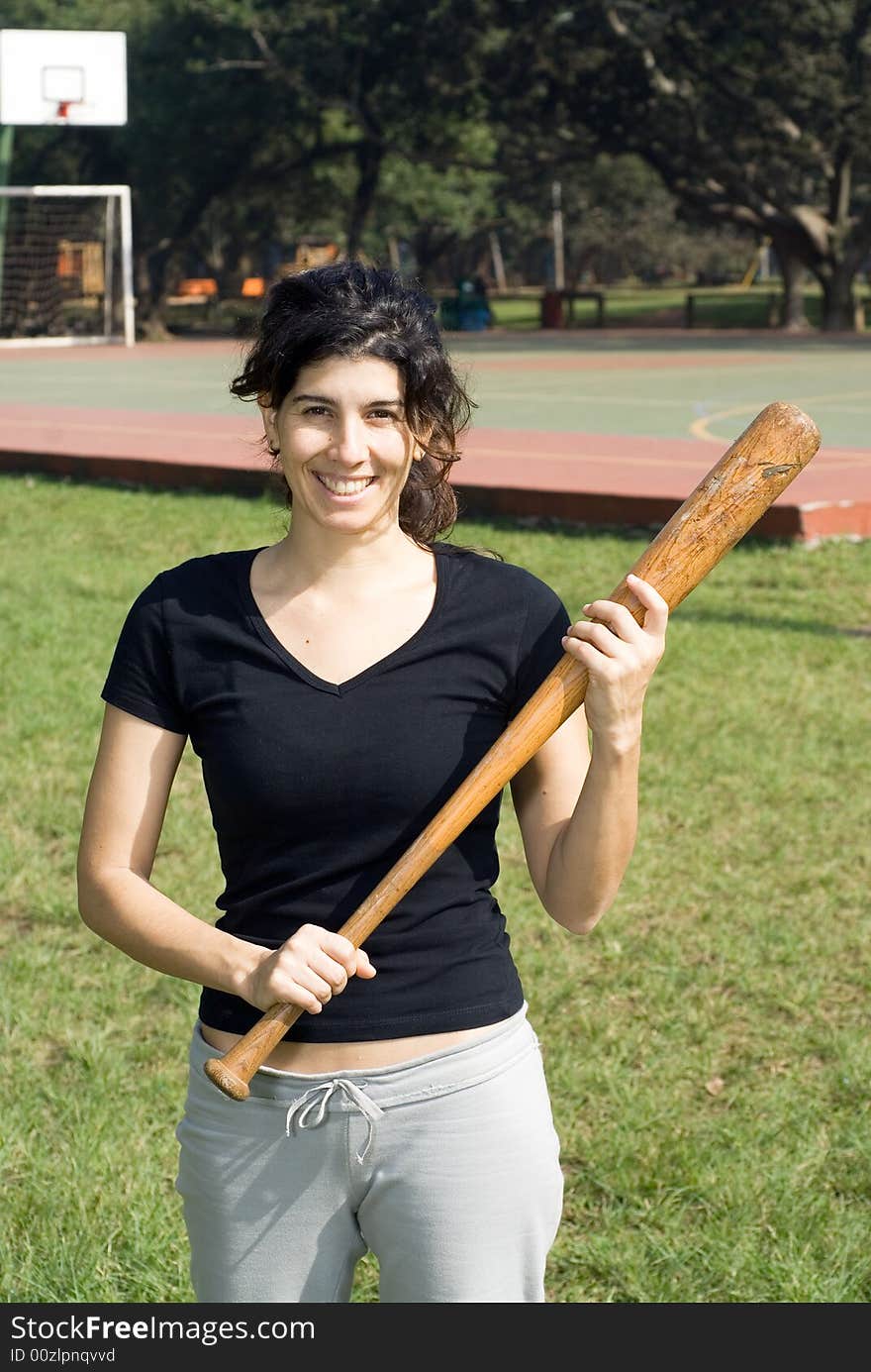 A smiling woman in a park is holding a baseball bat.  Vertically framed shot. A smiling woman in a park is holding a baseball bat.  Vertically framed shot.