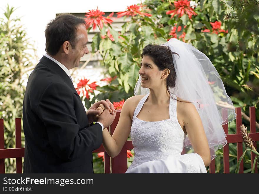 A newly married couple smile at each other while holding hands, red flowers predominant in background. - horizontally framed. A newly married couple smile at each other while holding hands, red flowers predominant in background. - horizontally framed