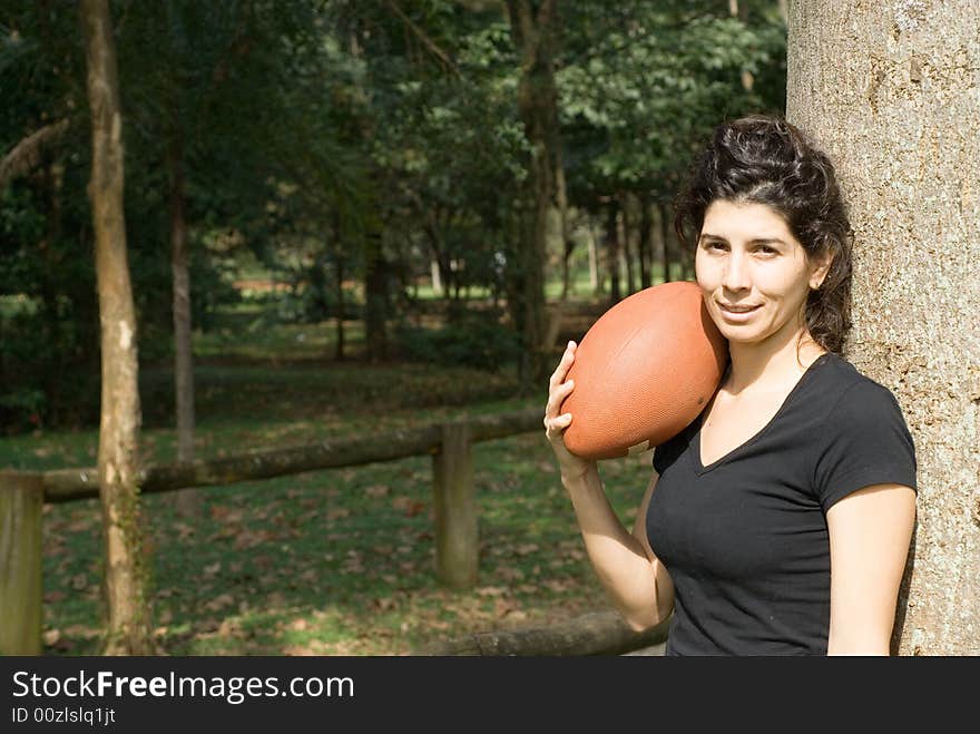 A young, attractive woman is standing next to a tree at the park. She is smiling and holding a football on her shoulder.Horizontally framed photo. A young, attractive woman is standing next to a tree at the park. She is smiling and holding a football on her shoulder.Horizontally framed photo.