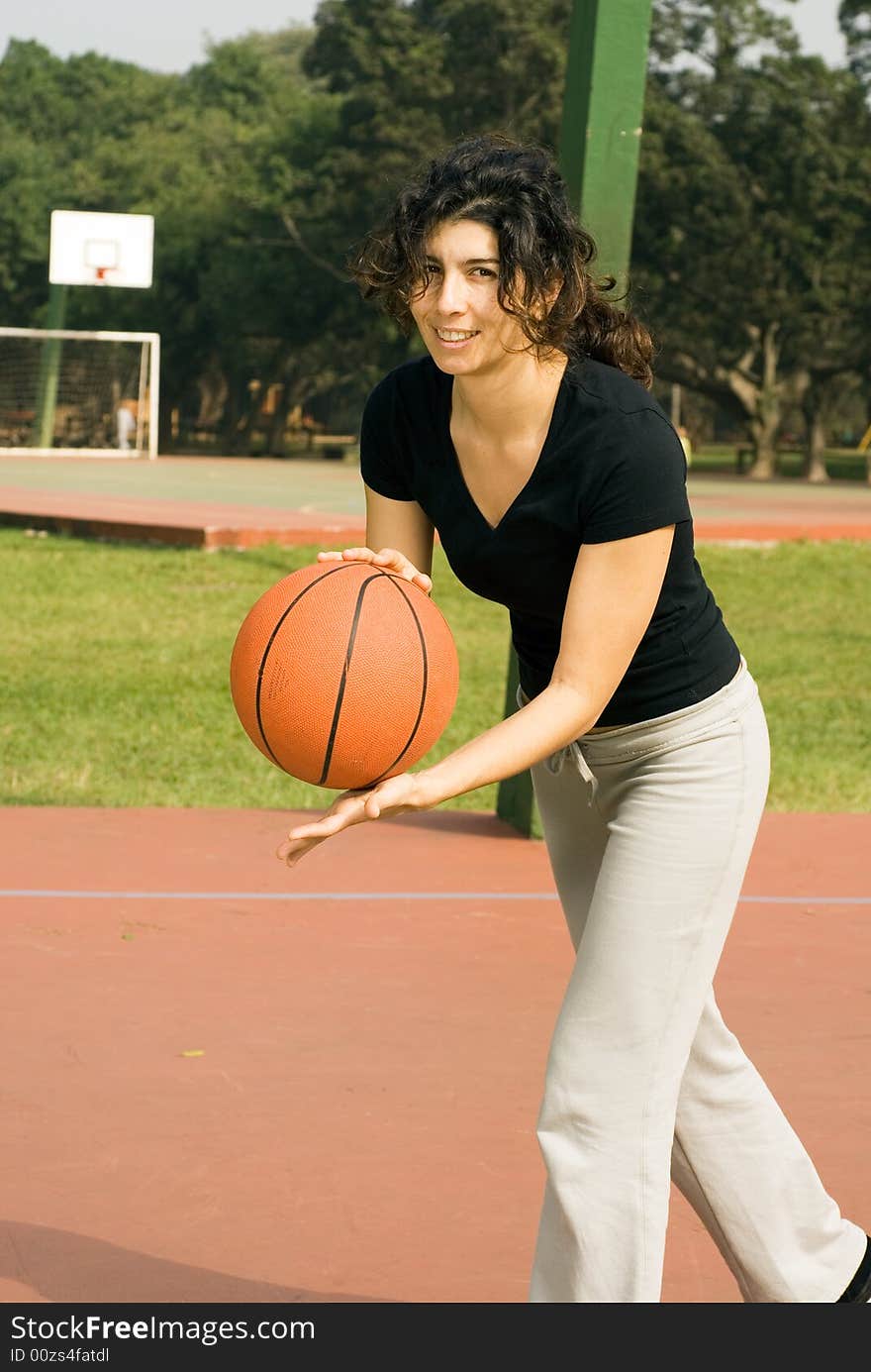 A woman is playing basketball on a park basketball court. She is smiling and looking at the camera. She looks like she is about to dribble the basketball. Vertically framed photo. A woman is playing basketball on a park basketball court. She is smiling and looking at the camera. She looks like she is about to dribble the basketball. Vertically framed photo.