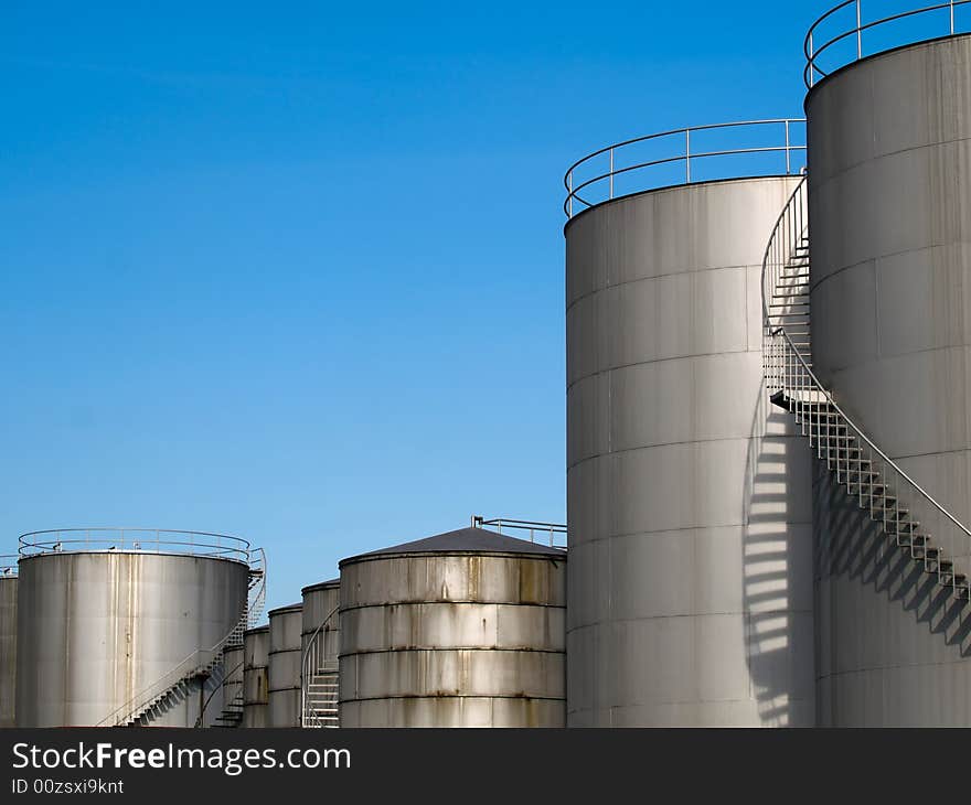 Row of cylindrical industrial storage tanks of various heights and sizes with spiral staircases on their sides leading to access on their roofs. Row of cylindrical industrial storage tanks of various heights and sizes with spiral staircases on their sides leading to access on their roofs