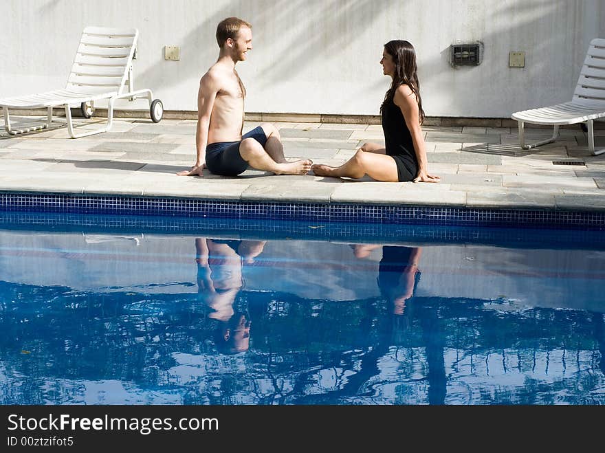 A couple, by the poolside, sit on the ground, stretching their groins, staring at each other, smiling - horizontally framed. A couple, by the poolside, sit on the ground, stretching their groins, staring at each other, smiling - horizontally framed