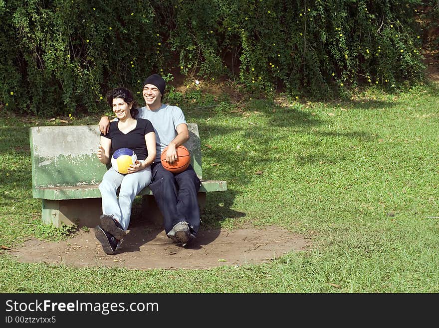 A couple is sitting on a park bench. They are both smiling and laughing and looking away from the camera and each other. The man is holding a basketball and the woman is holding a volleyball. Horizontally framed photo. A couple is sitting on a park bench. They are both smiling and laughing and looking away from the camera and each other. The man is holding a basketball and the woman is holding a volleyball. Horizontally framed photo.