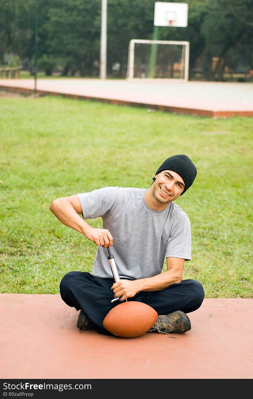 A man is sitting in a park. He is smiling, looking at the camera and pumping air into a football. Vertically framed photo. A man is sitting in a park. He is smiling, looking at the camera and pumping air into a football. Vertically framed photo.