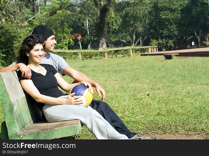 A couple is sitting on a park bench.  They are both smiling and looking away from the camera and each other.  The man has his arm around the woman and the woman is holding a volleyball.  Horizontally framed photo. A couple is sitting on a park bench.  They are both smiling and looking away from the camera and each other.  The man has his arm around the woman and the woman is holding a volleyball.  Horizontally framed photo.