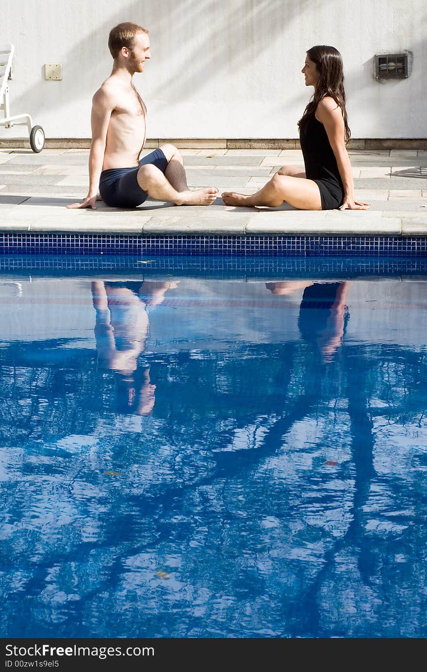 A couple, sitting by the poolside on the ground, stretching their groins, staring at each other, smiling - vertically framed. A couple, sitting by the poolside on the ground, stretching their groins, staring at each other, smiling - vertically framed