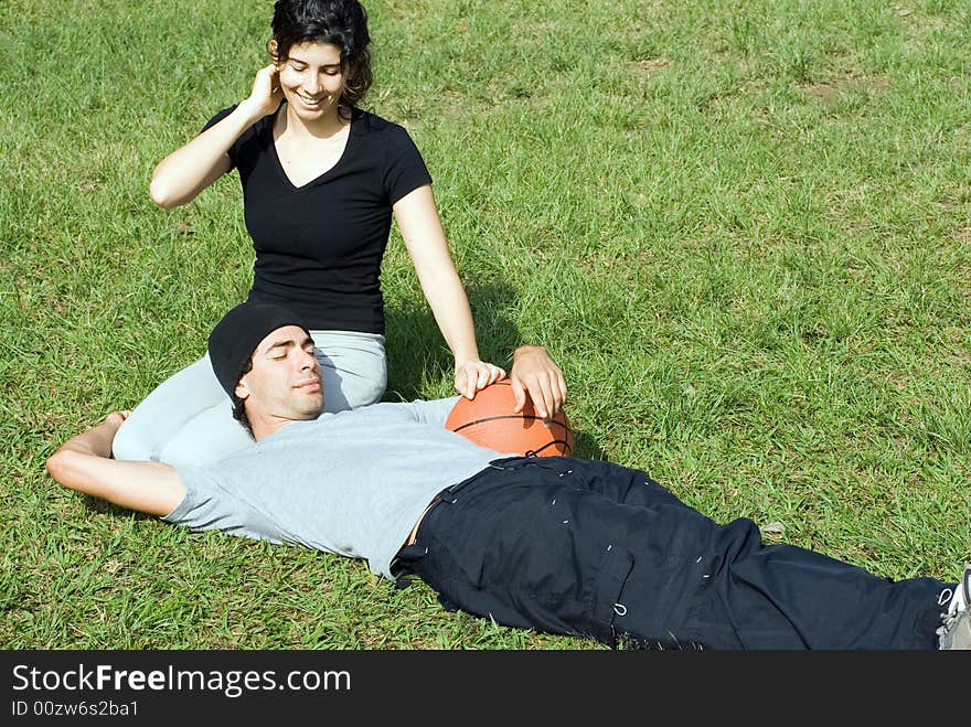 A couple is sitting on the grass in a park.  The woman is smiling and looking at the man and the man is resting on the womans lap.  The man is holding a basketball.  Horizontally framed photo. A couple is sitting on the grass in a park.  The woman is smiling and looking at the man and the man is resting on the womans lap.  The man is holding a basketball.  Horizontally framed photo.