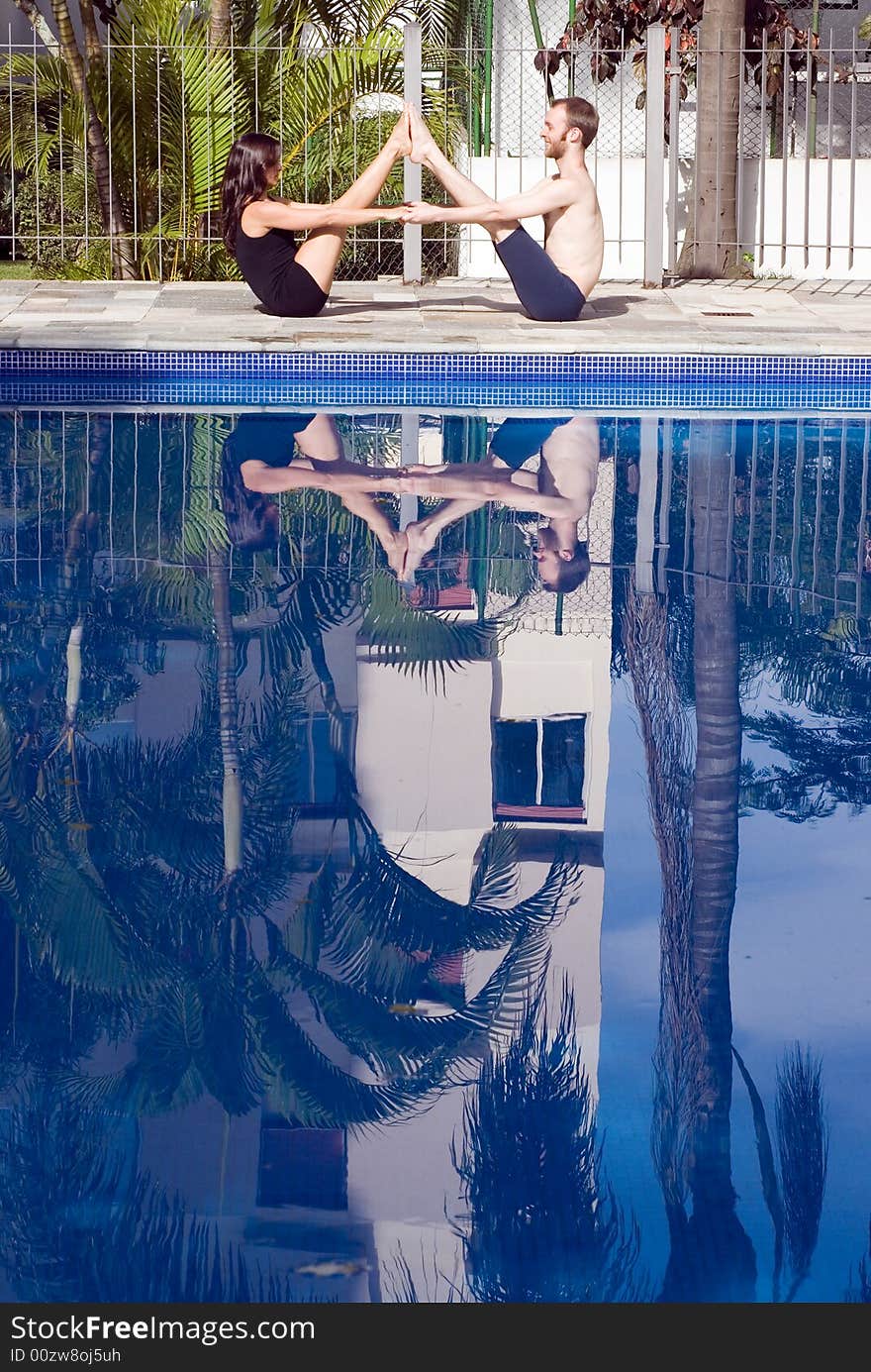 A couple, with their feet touching and hands linked, stretch with each other next to the pool - vertically framed. A couple, with their feet touching and hands linked, stretch with each other next to the pool - vertically framed