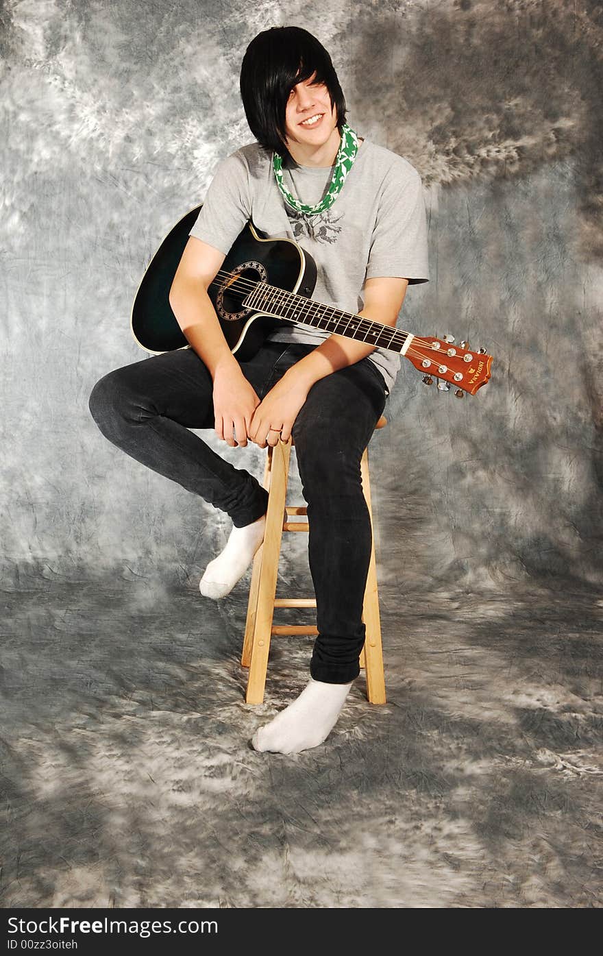 An boy in black jeans sitting on a chair with an green guitar for a black 
and white background and smiling in the camera. An boy in black jeans sitting on a chair with an green guitar for a black 
and white background and smiling in the camera.