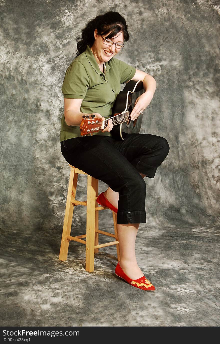 An woman in black pants sitting on a chair with an green guitar for a black and white background and smiling in the camera. An woman in black pants sitting on a chair with an green guitar for a black and white background and smiling in the camera.