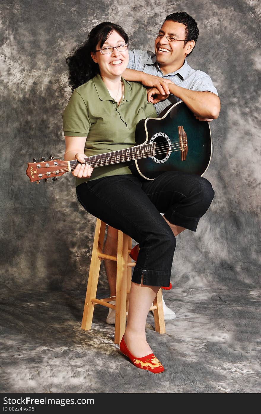 An woman in black pants sitting on a chair with an green guitar for a black 
and white background and smiling in the camera, with her husband. An woman in black pants sitting on a chair with an green guitar for a black 
and white background and smiling in the camera, with her husband.