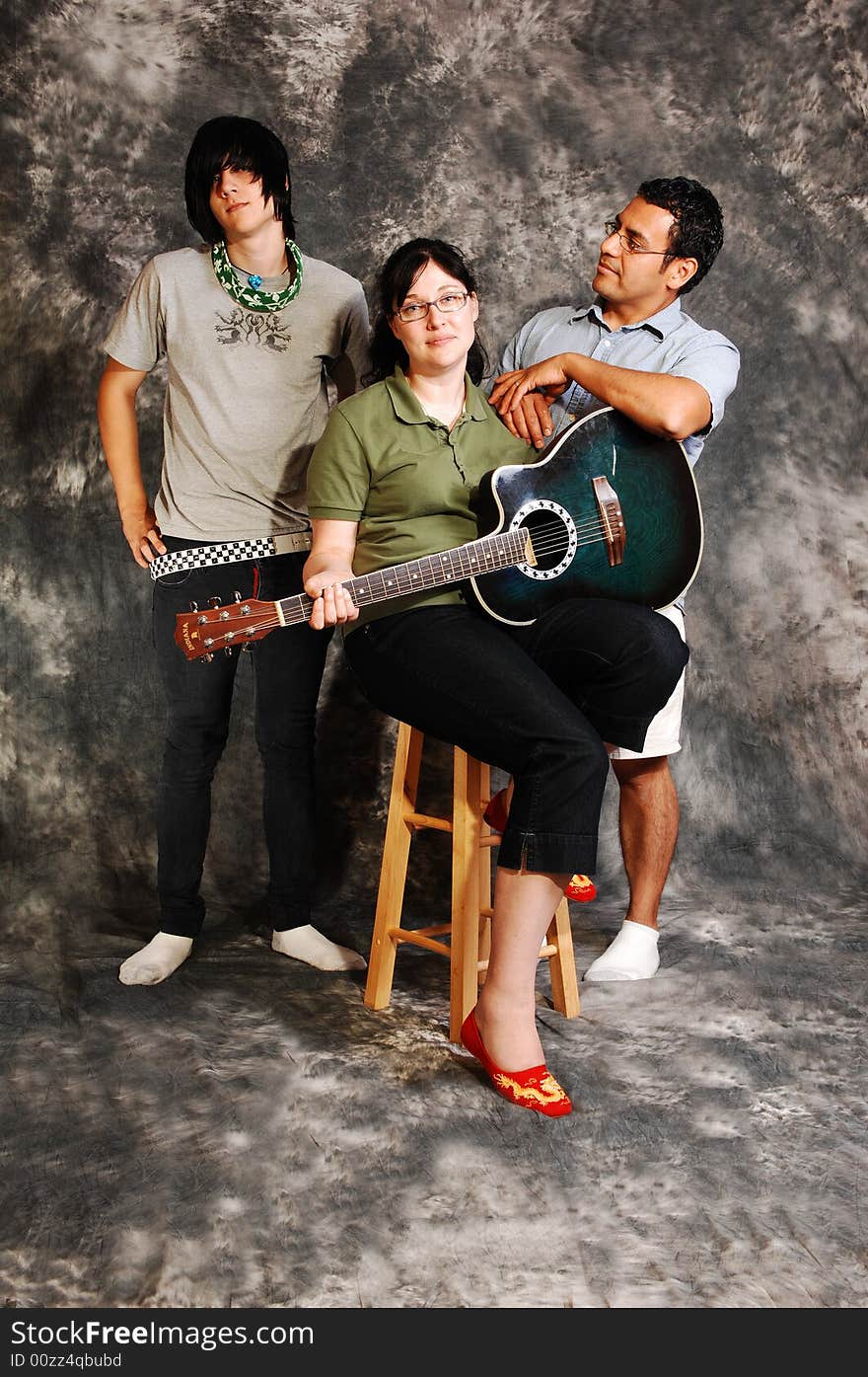 An woman in black pants sitting on a chair with an green guitar for a black 
and white background and smiling in the camera, with her husband and son. An woman in black pants sitting on a chair with an green guitar for a black 
and white background and smiling in the camera, with her husband and son.