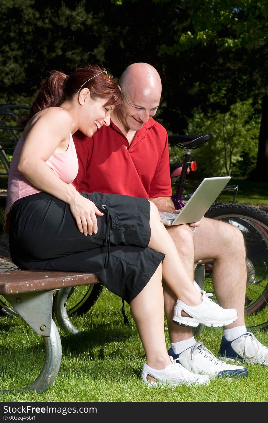 Couple on a park bench laugh as they look at a laptop computer, their bikes are in the background. Vertically framed photograph. Couple on a park bench laugh as they look at a laptop computer, their bikes are in the background. Vertically framed photograph