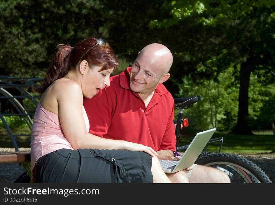 Couple on a park bench looking at a laptop computer, he is smiling at her and she looks surprised. Their bikes are in the background. Horizontally framed photograph. Couple on a park bench looking at a laptop computer, he is smiling at her and she looks surprised. Their bikes are in the background. Horizontally framed photograph