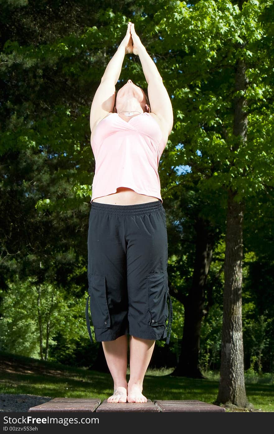 Woman standing on picnic table in the park. Hands positioned above head and in prayer formation. Head and body leaning back, feet maintianing balance. Vertically framed shot. Woman standing on picnic table in the park. Hands positioned above head and in prayer formation. Head and body leaning back, feet maintianing balance. Vertically framed shot.