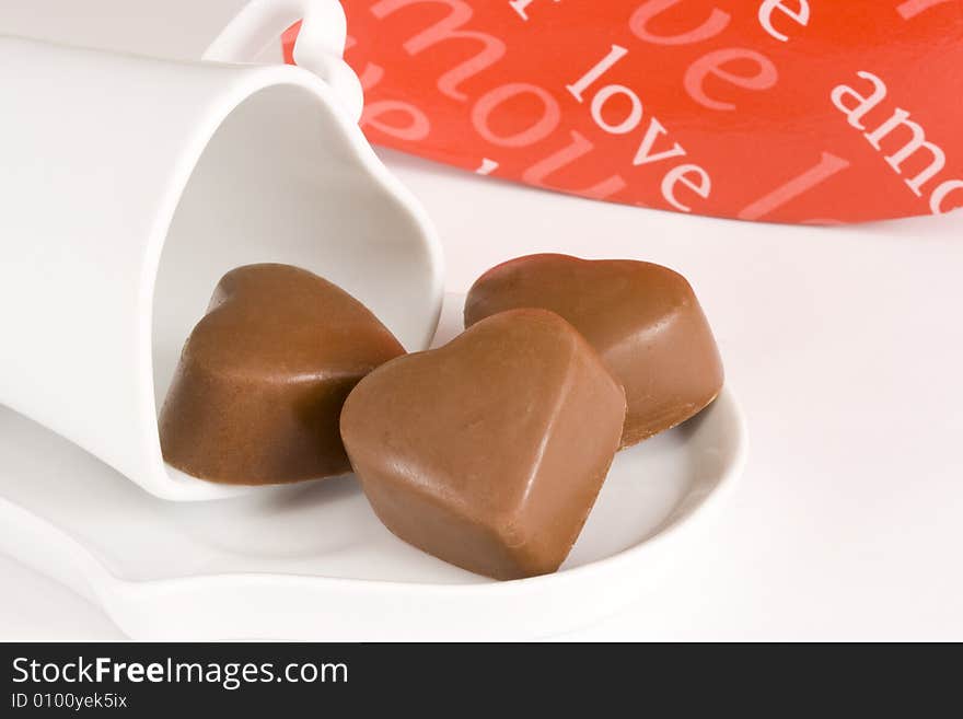 Three brown heart shaped chocolates placed on a heart shaped cup and causer with red box in background, all on white. Three brown heart shaped chocolates placed on a heart shaped cup and causer with red box in background, all on white