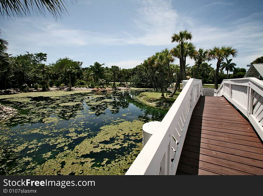 A photo of a white bridge crossing over a man made pond. A photo of a white bridge crossing over a man made pond.