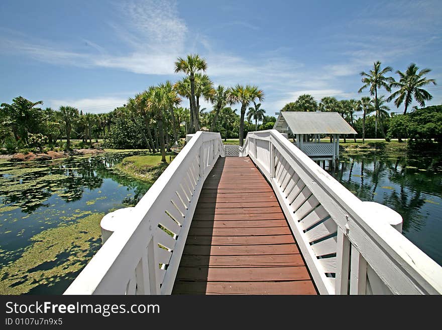 A photo of a white bridge crossing over a man made pond to an island. A photo of a white bridge crossing over a man made pond to an island.