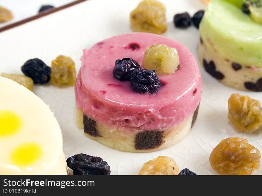 A close up of a red fruits petit four with blueberry and grape on top, surrounded by others petit fours and raisins. Main focus on the central petit four.