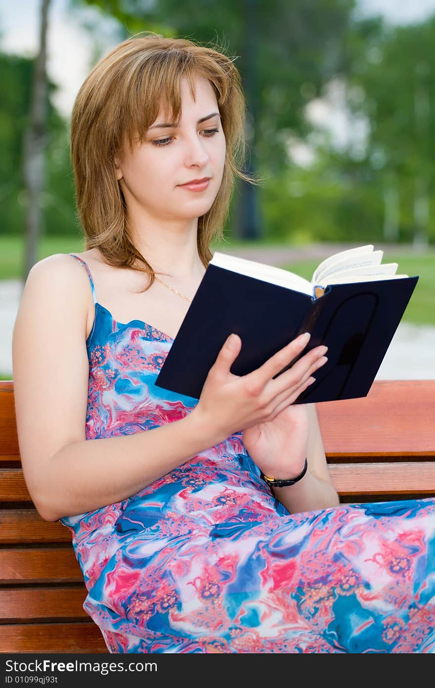 Attractive young girl reads the book sitting on a bench in park