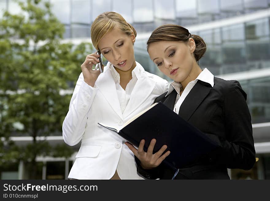 Two attractive businesswomen talking on the phone and holding a notebook outdoors. Two attractive businesswomen talking on the phone and holding a notebook outdoors