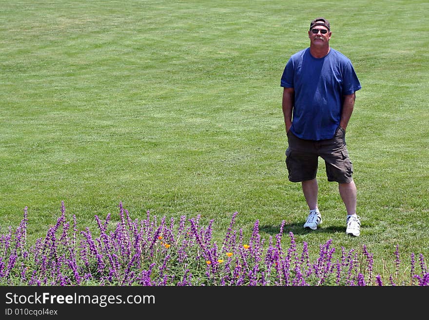 A man standing on a green lawn with hands in his pockets, tall purple flowers in foreground, room for copyspace on left. A man standing on a green lawn with hands in his pockets, tall purple flowers in foreground, room for copyspace on left