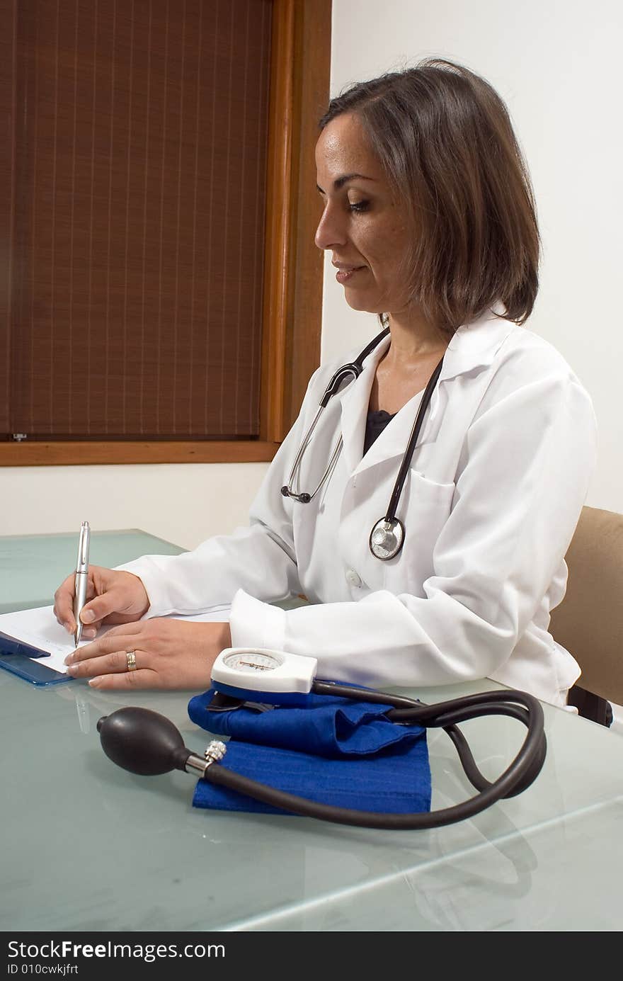 A female doctor, sits at her desk with her blood pressure tester on it, writing in her clipboard. - vertically framed. A female doctor, sits at her desk with her blood pressure tester on it, writing in her clipboard. - vertically framed