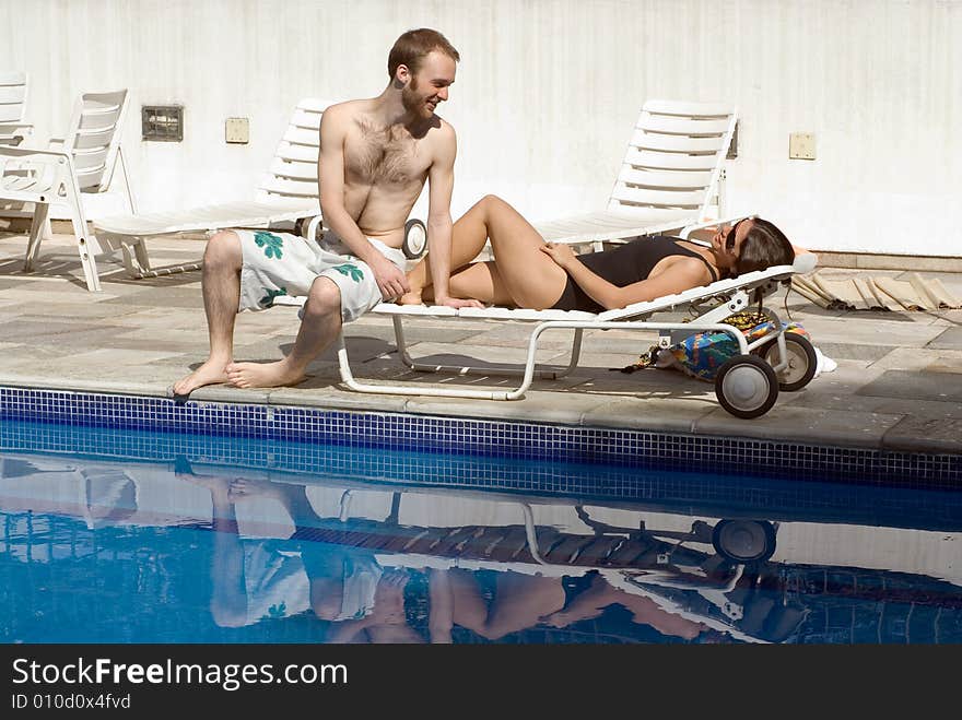 A young couple, share a pool chair, the woman lying, while the man sits near her feet. - horizontally framed. A young couple, share a pool chair, the woman lying, while the man sits near her feet. - horizontally framed