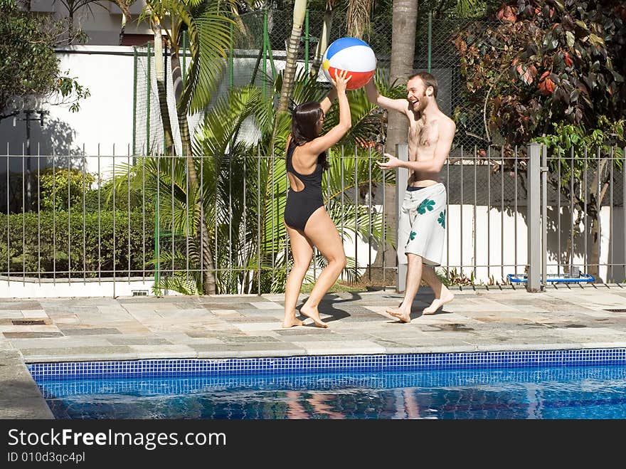 A man and a woman are playing together next to a pool.  They are smiling and laughing and looking at each other.  They are holding a beachball.  Horizontally framed photo. A man and a woman are playing together next to a pool.  They are smiling and laughing and looking at each other.  They are holding a beachball.  Horizontally framed photo.