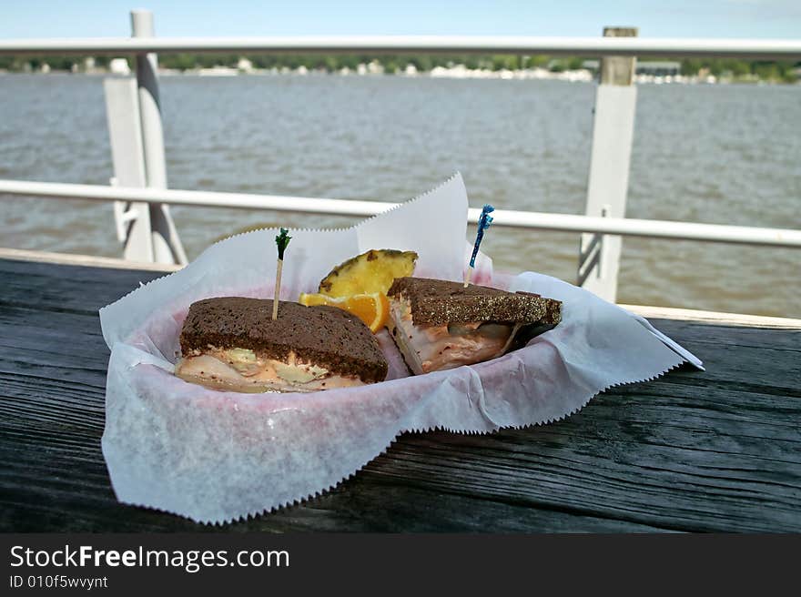 A sandwich in a basket on a rough table with a lake and shore line in the background. A sandwich in a basket on a rough table with a lake and shore line in the background