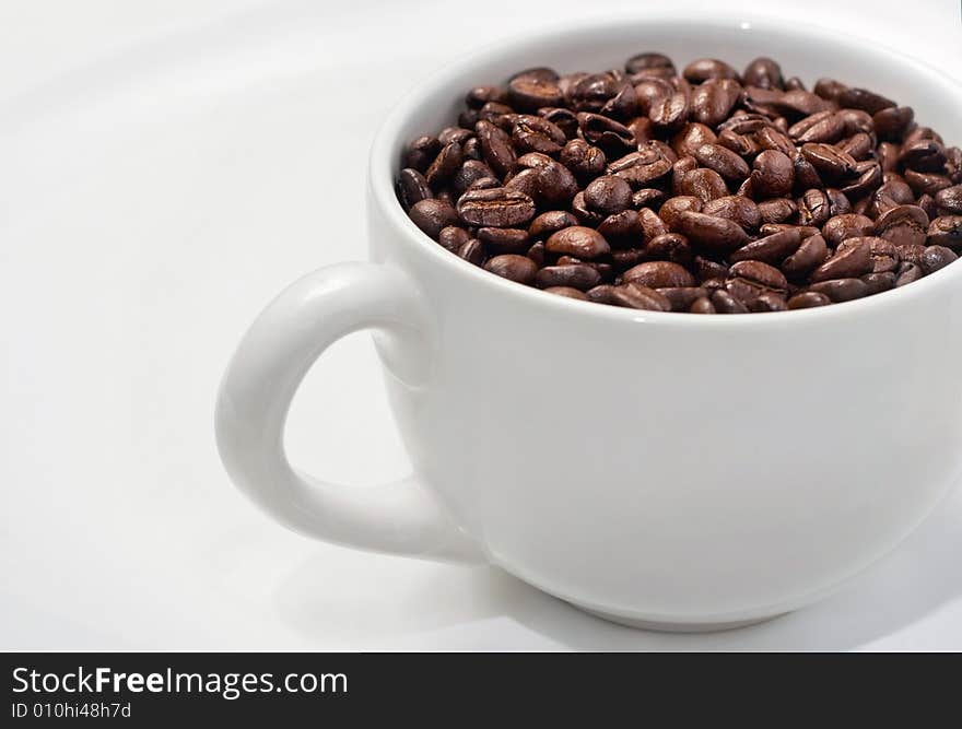 Rich brown beans fill up a classic white coffee mug sitting on large white saucer. Rich brown beans fill up a classic white coffee mug sitting on large white saucer