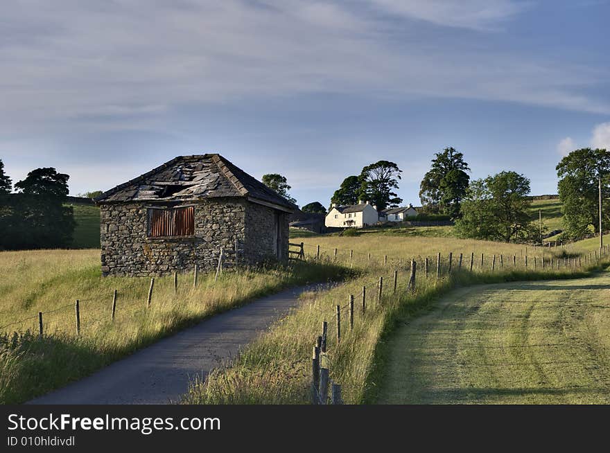 Small ruined barn on a rural road leading to a white farmhouse in the English Lake District. Small ruined barn on a rural road leading to a white farmhouse in the English Lake District