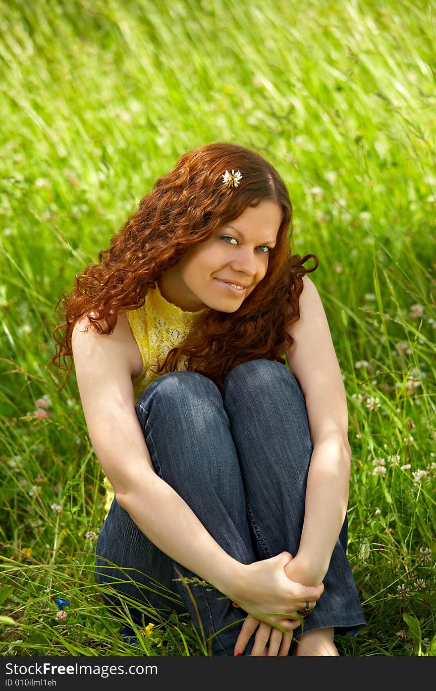 The beautiful young girl sits on a background of a green lawn and smiles. The beautiful young girl sits on a background of a green lawn and smiles