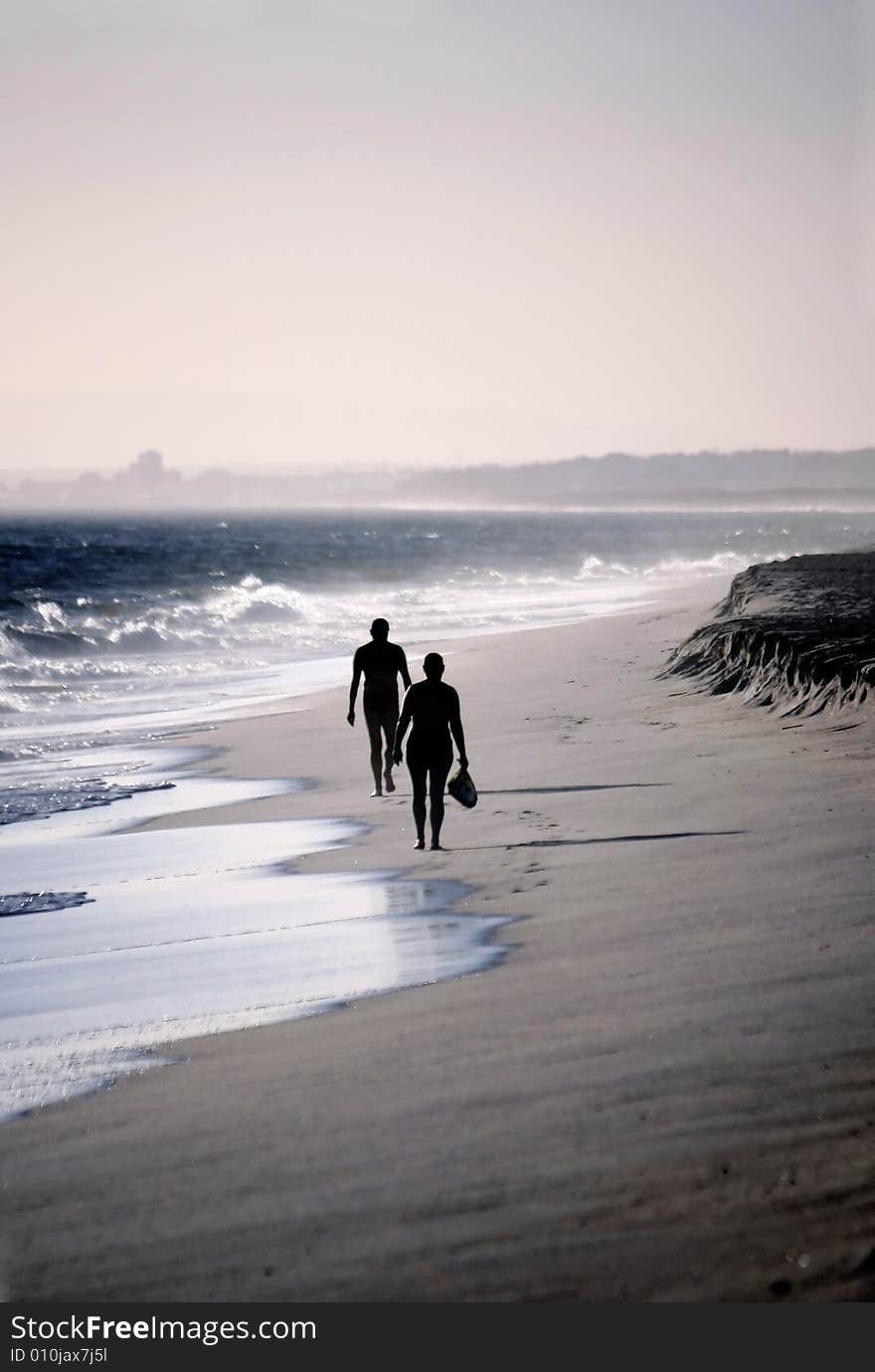 Two lonely figures walking down the beach shoreline on a windy day.