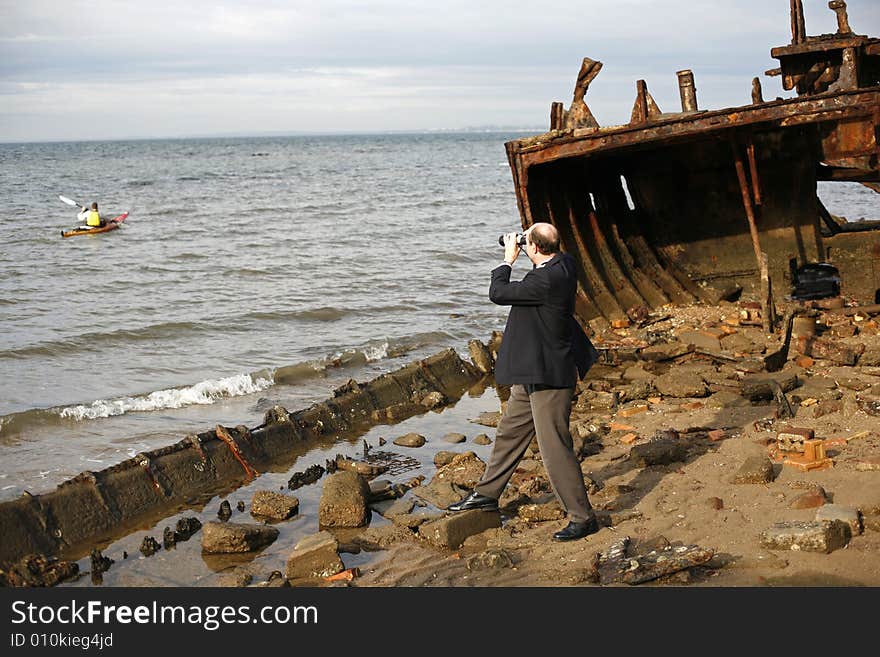 Businessman in the rusting hull of shipwreck, with binoculars looking out to the horizon where canoeist is, asking the question is their an escape from a sinking business. Businessman in the rusting hull of shipwreck, with binoculars looking out to the horizon where canoeist is, asking the question is their an escape from a sinking business